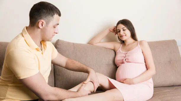 Happy pregnant family. Husband massaging his pregnant wife legs and sitting together on couch in their home.
